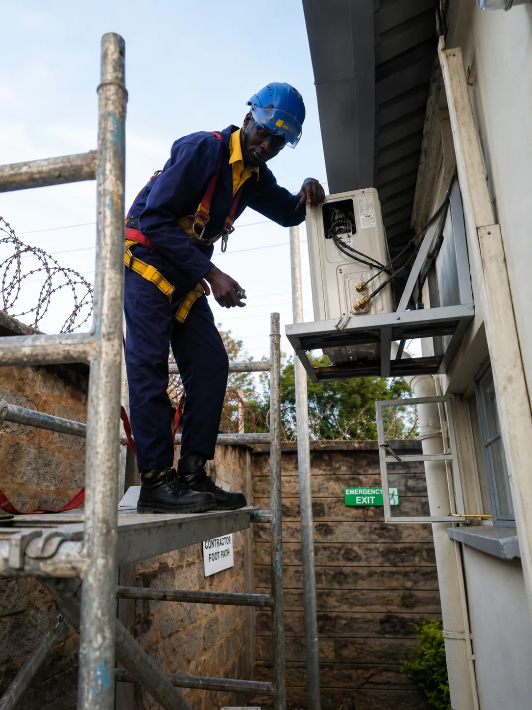Technician on scaffolding with a fall-arrest harness working on a wall-mounted outdoor unit